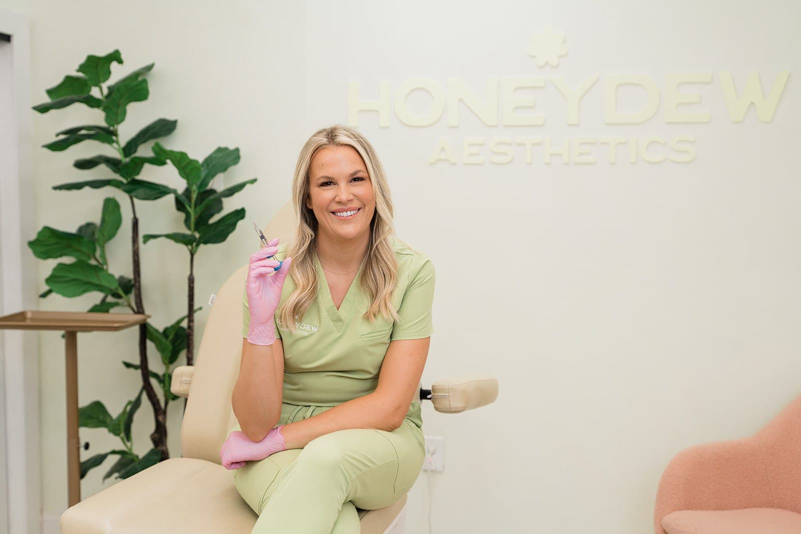 Taryn, RN, smiling in a treatment chair holding a syringe beneath the Honeydew Aesthetics wall sign